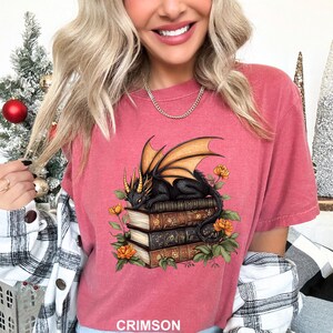 A woman wearing a pink t-shirt with a graphic of a dragon and books, smiling and holding a red ornament.