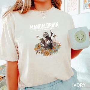 A person wearing a white t-shirt with a graphic design of a character from the Mandalorian television series, holding a coffee mug with a smiley face on it.