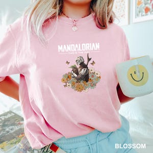 A person wearing a pink t-shirt with a graphic of a character from the Mandalorian TV series, holding a coffee mug with a smiley face.