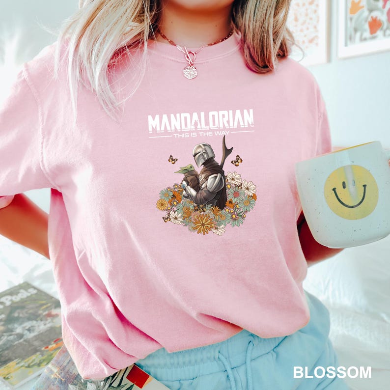 A person wearing a pink t-shirt with a graphic of a character from the Mandalorian TV series, holding a coffee mug with a smiley face.