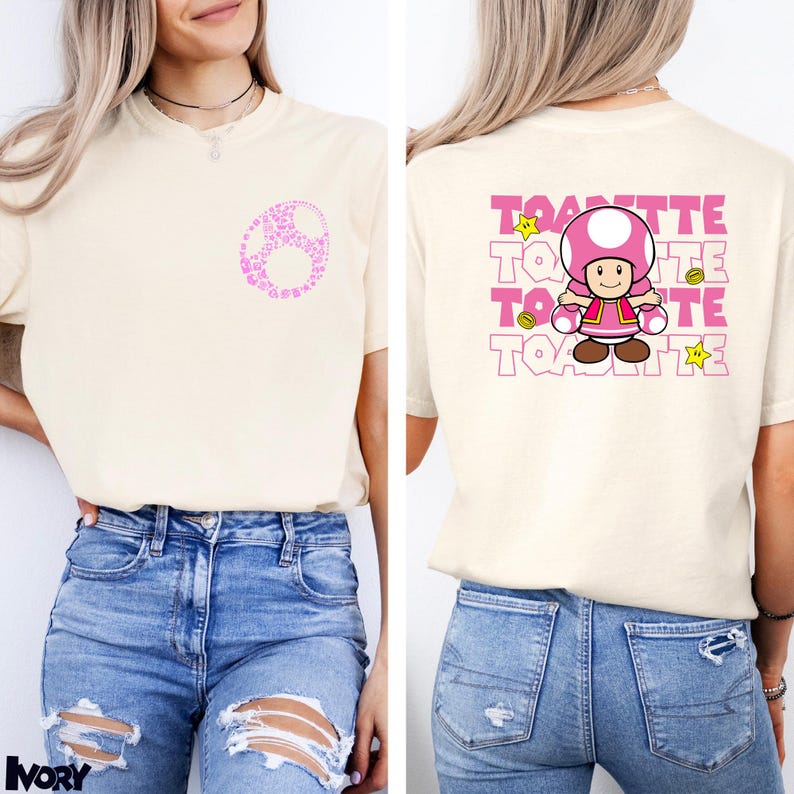 May include: An ivory t-shirt featuring a small pink circular graphic on the front left chest. The back displays a large graphic of the character Toadette, surrounded by the repeated text TOADETTE in pink, along with yellow star and coin icons.