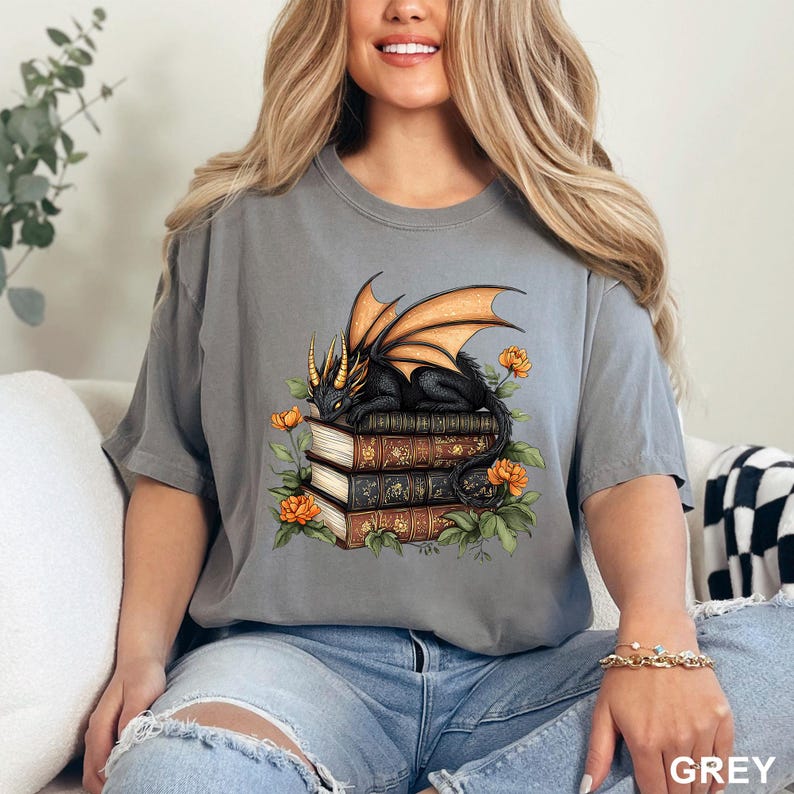 A woman wearing a gray t-shirt with a graphic design of a dragon perched on a stack of books, surrounded by flowers.