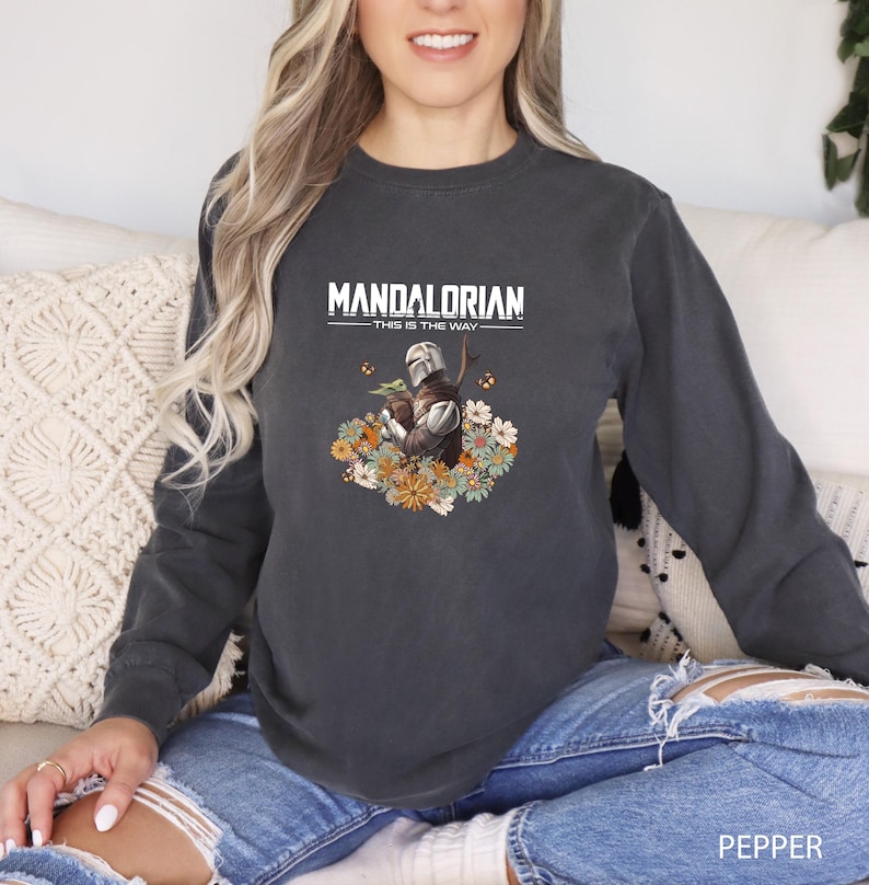 A woman wearing a gray sweatshirt with a graphic of a character from the Star Wars series, specifically Mandalorian, sitting on a couch.