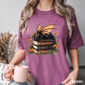 A woman wearing a purple t-shirt with a graphic design of a dragon perched on a stack of books, surrounded by flowers.