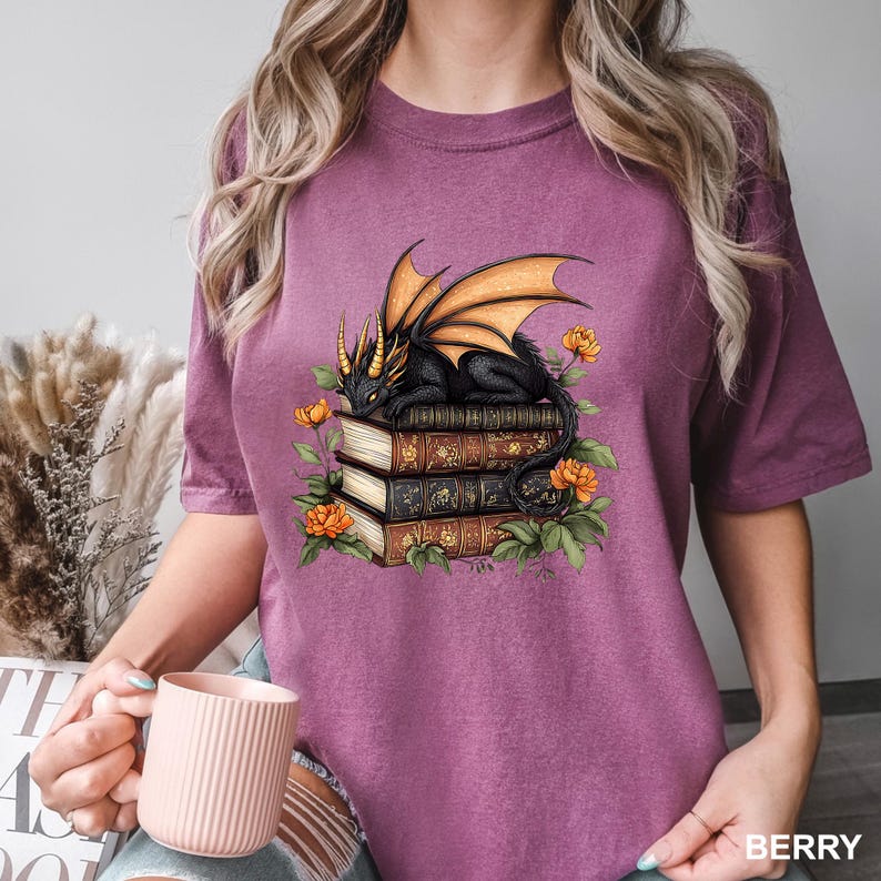 A woman wearing a purple t-shirt with a graphic design of a dragon perched on a stack of books, surrounded by flowers.