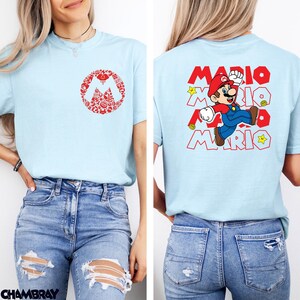 May include: A light blue t-shirt featuring a red circular graphic on the front chest composed of small Mario-themed icons. The back displays a large graphic of Mario jumping with the text MARIO repeated in red outline font and yellow stars.