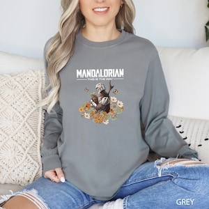 A woman wearing a grey long-sleeved shirt with a graphic of a character from the Mandalorian television series, sitting on a couch.