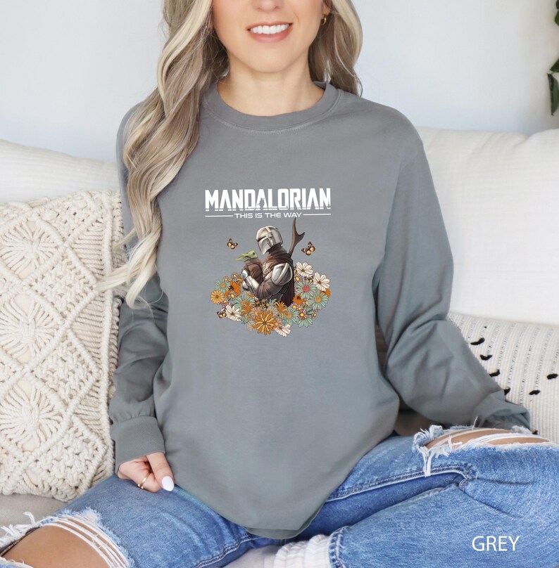 A woman wearing a grey long-sleeved shirt with a graphic of a character from the Mandalorian television series, sitting on a couch.