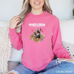 A woman wearing a pink long-sleeved shirt with a graphic design of a character from the Mandalorian television series.