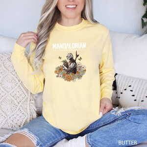A woman wearing a yellow sweatshirt with a Mandalorian graphic and text, sitting on a couch.
