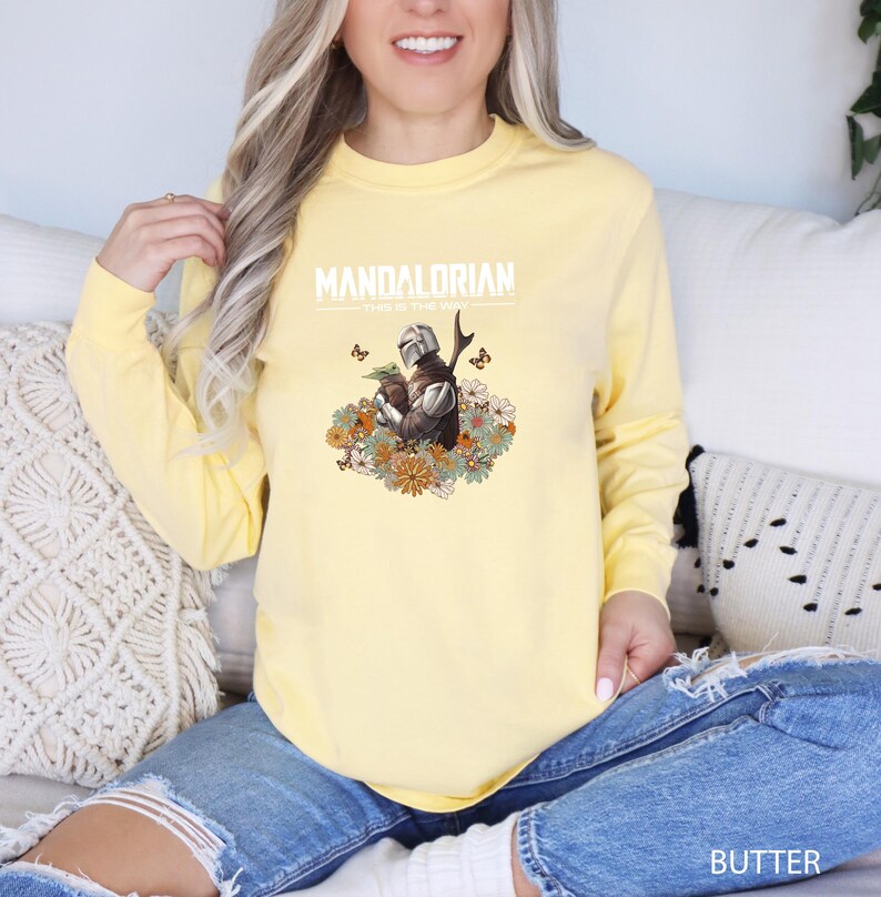 A woman wearing a yellow sweatshirt with a Mandalorian graphic and text, sitting on a couch.