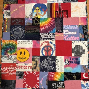 May include: A rectangular patchwork quilt constructed from various graphic t-shirt squares featuring diverse prints, including text, logos, tie-dye patterns, and illustrations, all stitched together with a floral patterned border trim.