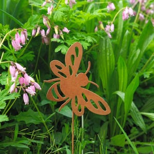 Peut inclure: Un piquet de jardin en métal brun rouillé en forme d'abeille avec des détails découpés. L'abeille est positionnée dans un jardin avec un feuillage vert et des fleurs roses. Le piquet est conçu pour être planté dans le sol comme élément décoratif.
