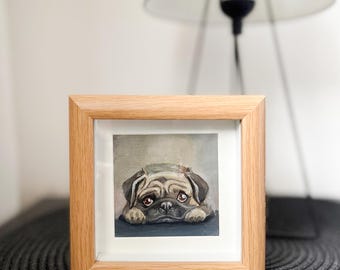 Puede incluir: Una obra de arte cuadrada enmarcada que presenta un perro pug con una expresión triste. El pug está pintado en tonos marrones, beige y grises, con ojos grandes y expresivos. La obra de arte está enmarcada en un marco de madera clara. Una lámpara blanca está en el fondo.