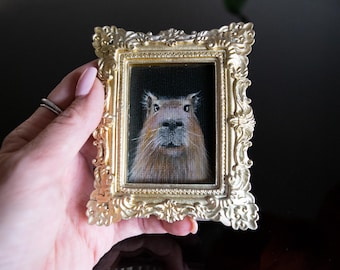 Puede incluir: Un pequeño marco dorado adornado con una pintura de un capibara en su interior. El capibara está mirando directamente al espectador con una expresión curiosa.