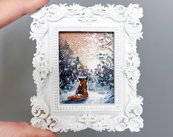 Puede incluir: Pequeña pintura de un zorro en un bosque nevado, enmarcada en un marco blanco ornamentado. La obra presenta un zorro marrón, árboles cubiertos de nieve y un cielo de tonos cálidos. El marco tiene un diseño detallado de volutas.