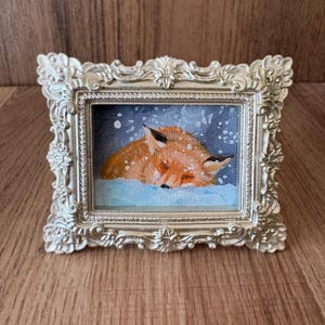 Puede incluir: Peque&ntilde;a pintura enmarcada que representa un zorro durmiendo en la nieve. El zorro, en tonos naranja y marr&oacute;n, descansa sobre un fondo nevado azul. El marco plateado, con detalles ornamentales, realza la obra.