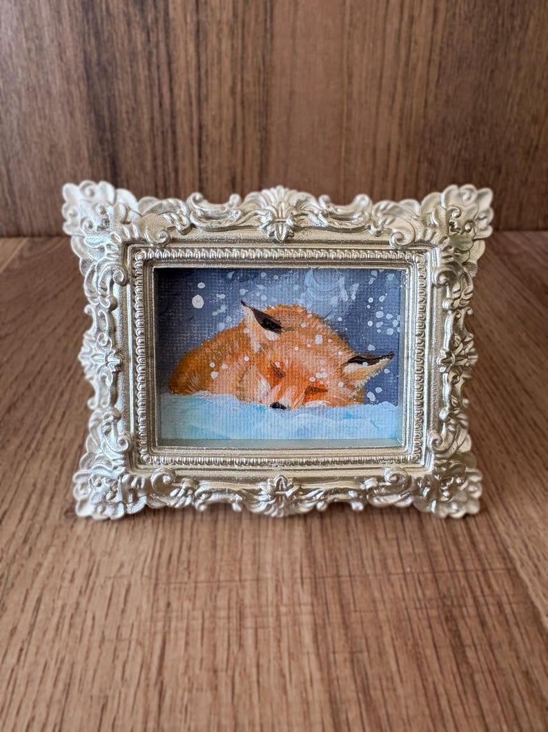 Puede incluir: Peque&ntilde;a pintura enmarcada que representa un zorro durmiendo en la nieve. El zorro, en tonos naranja y marr&oacute;n, descansa sobre un fondo nevado azul. El marco plateado, con detalles ornamentales, realza la obra.