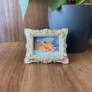 Puede incluir: Una peque&ntilde;a pintura enmarcada con dos zorros naranjas durmiendo en la nieve. La pintura est&aacute; en un marco dorado ornamentado. El fondo es azul nevado. El marco est&aacute; sobre una superficie de madera.