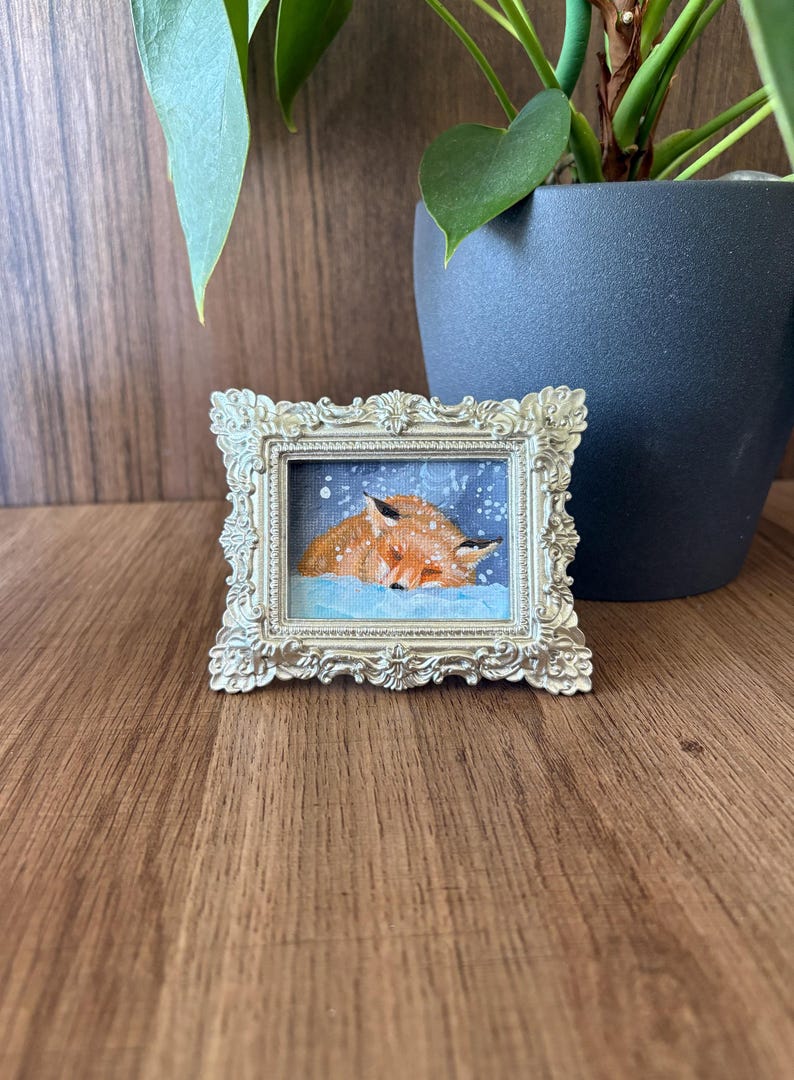 Puede incluir: Una peque&ntilde;a pintura enmarcada con dos zorros naranjas durmiendo en la nieve. La pintura est&aacute; en un marco dorado ornamentado. El fondo es azul nevado. El marco est&aacute; sobre una superficie de madera.