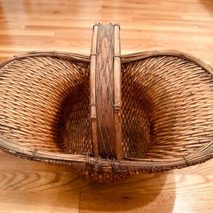 以下が含まれることがあります： 木と編まれたストリップで作られたハンドルが付いた、大きな籐のバスケット。バスケットは自然素材で作られており、薄い茶色をしています。