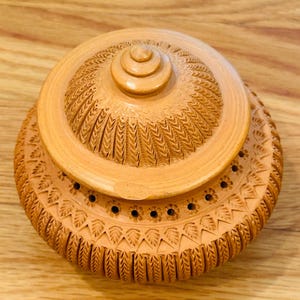 以下が含まれることがあります： テラコッタ色の蓋付きセラミックポット。複雑な彫刻模様が施されています。蓋には同心円と中央のノブがあります。ポットの本体には、一連の小さな穴と、詳細な花と葉のデザインがあります。