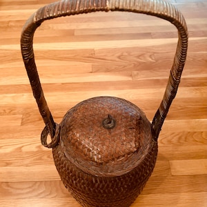 Può includere: Un cesto intrecciato marrone con un coperchio e un manico lungo. Il cesto è fatto di materiali naturali e ha un aspetto rustico.