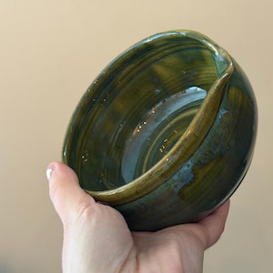 May include: A handmade ceramic bowl with a unique, asymmetrical design. The bowl features a blend of green and gold hues, with a glossy finish. The interior has a spiral pattern, and the rim is slightly raised.