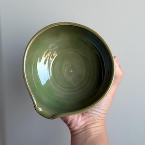 May include: A round, olive green ceramic bowl with a smooth, glossy interior finish and a small, integrated pouring spout on one side. The bowl features subtle, circular wheel-thrown markings visible at the base of the interior basin.