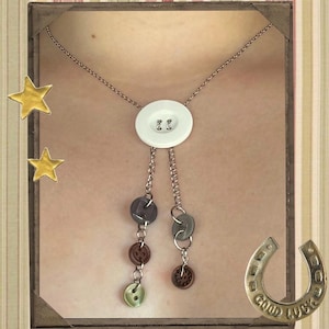 May include: A handmade necklace featuring a large white button pendant with a chain. The necklace has dangling chains with various colored buttons. The background includes gold stars and a horseshoe charm with the words "Good Luck".