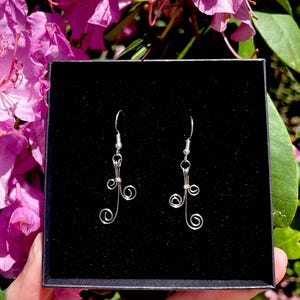 May include: A pair of silver-toned wire earrings with a delicate, swirling design. The earrings are displayed in a black box against a backdrop of vibrant pink flowers and green foliage. The earrings have a hook closure.