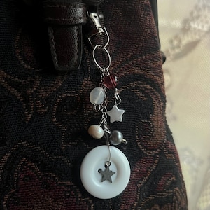 May include: A silver keychain with a decorative charm. The charm features a white circular pendant with a star, along with dangling beads and star-shaped charms. The keychain is attached to a dark brown leather bag.