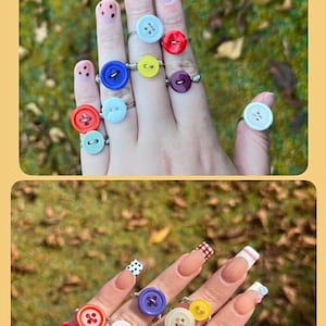 Peut inclure: Anneaux en forme de boutons de différentes couleurs, dont le rouge, le bleu, le jaune et le violet. Les anneaux sont montés sur des bagues en argent et présentés sur une main aux ongles manucurés. L'arrière-plan est une vue floue d'herbe verte.