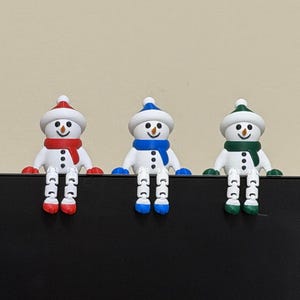 May include: Three decorative snowman figurines with red, blue, and green accents. Each snowman has a matching hat and scarf, with black button eyes and a carrot nose. The snowmen are sitting on a black surface.