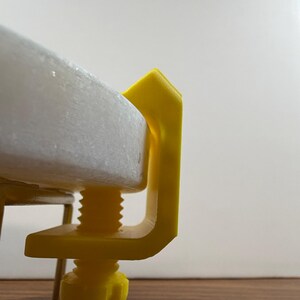 May include: A bright yellow C-clamp made of plastic, featuring a threaded screw mechanism with a knob at the bottom. The clamp is securely attached to the edge of a thick, circular white stone disc, holding it firmly in place against a metal frame.