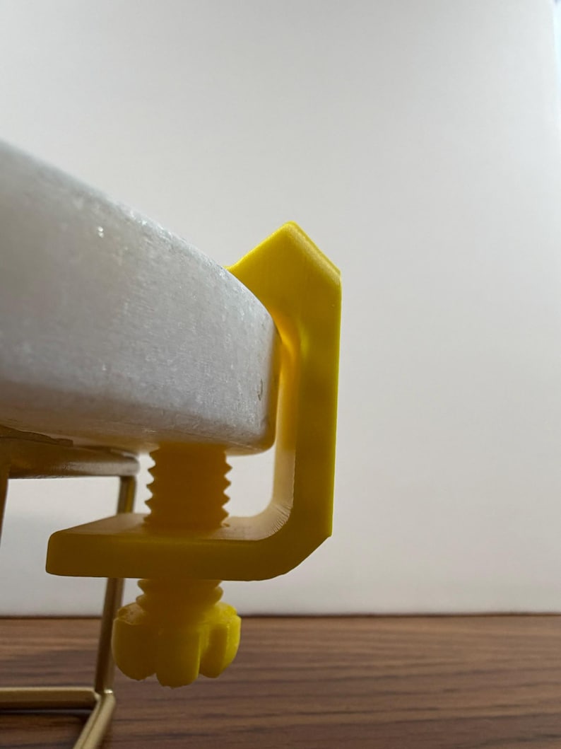 May include: A bright yellow C-clamp made of plastic, featuring a threaded screw mechanism with a knob at the bottom. The clamp is securely attached to the edge of a thick, circular white stone disc, holding it firmly in place against a metal frame.