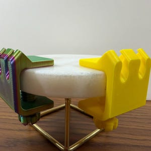 May include: Two C-shaped clamps attached to a circular white stone disc. One clamp is a solid bright yellow, while the other features a gradient of green, purple, and blue. Each clamp has a threaded screw mechanism underneath for tightening.