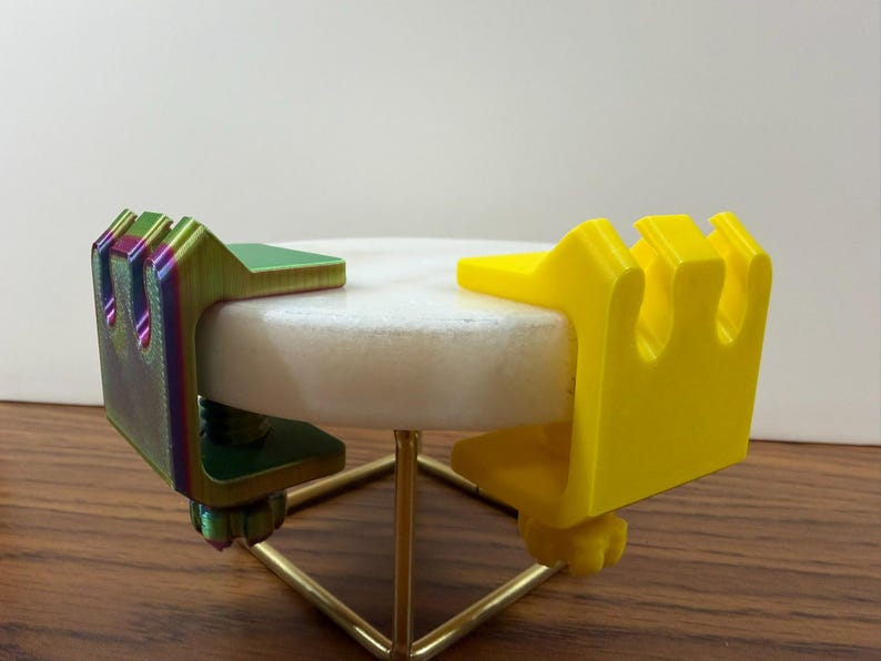 May include: Two C-shaped clamps attached to a circular white stone disc. One clamp is a solid bright yellow, while the other features a gradient of green, purple, and blue. Each clamp has a threaded screw mechanism underneath for tightening.
