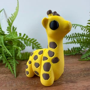 May include: A small, yellow giraffe figurine with dark brown spots and large, solid black eyes. The giraffe has a smooth, textured surface and is depicted in a standing position with its head turned slightly to the right, showing its ears and ossicones.