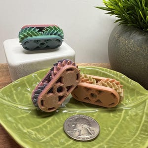 May include: Three small, colorful, 3D printed, gear-shaped toys. The toys are made of plastic and have a gradient color scheme. The toys are on a green leaf-shaped dish and a white square platform.