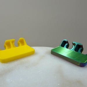 May include: Two small, rectangular plastic clips with three distinct, curved prongs extending from one side. One clip is a solid, bright yellow, while the other features a metallic, iridescent finish that shifts between green, blue, and purple hues.