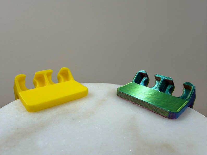 May include: Two small, rectangular plastic clips with three distinct, curved prongs extending from one side. One clip is a solid, bright yellow, while the other features a metallic, iridescent finish that shifts between green, blue, and purple hues.