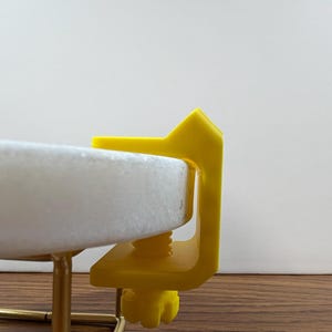 May include: A bright yellow C-clamp with a threaded screw mechanism attached to the edge of a circular white stone disc. The clamp features a flat top surface and a textured adjustment knob at the bottom, securing the disc to a thin gold metal frame.