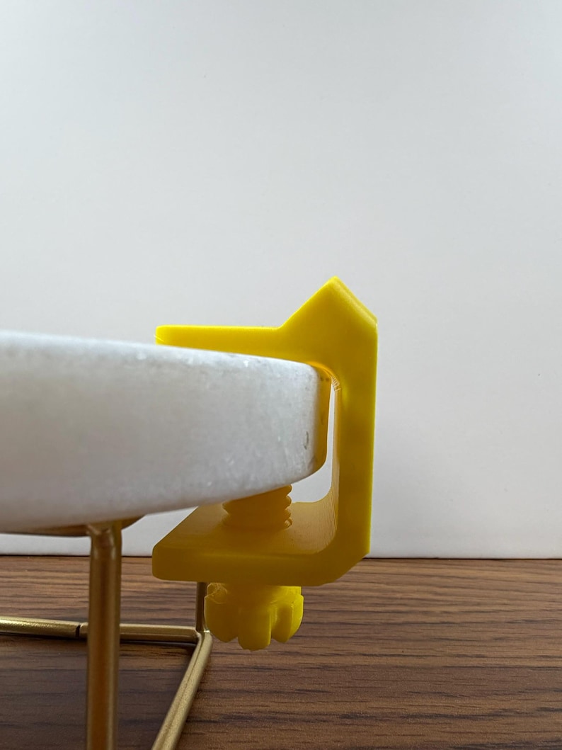 May include: A bright yellow C-clamp with a threaded screw mechanism attached to the edge of a circular white stone disc. The clamp features a flat top surface and a textured adjustment knob at the bottom, securing the disc to a thin gold metal frame.
