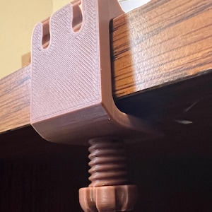May include: A brown 3D-printed C-clamp featuring a U-shaped body with two vertical slots at the top and a threaded screw mechanism with a circular knob at the bottom, designed to secure onto the edge of a flat surface for mounting purposes.