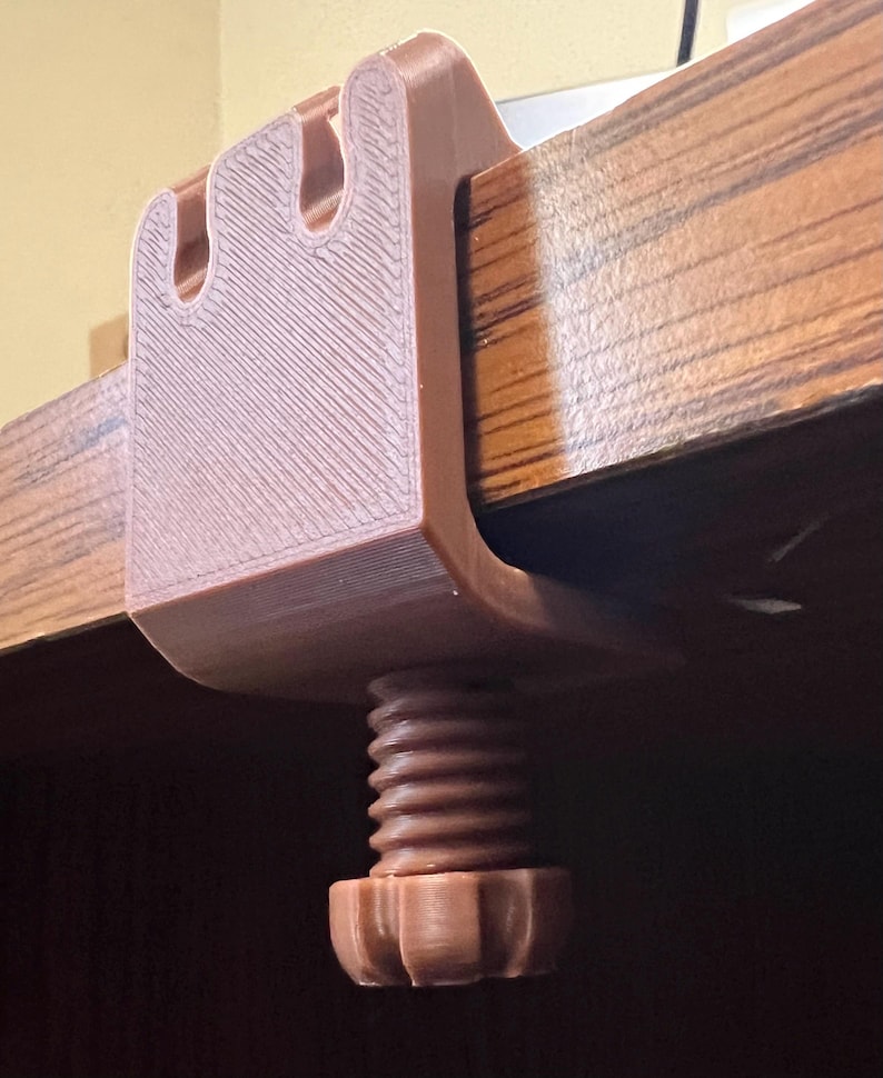 May include: A brown 3D-printed C-clamp featuring a U-shaped body with two vertical slots at the top and a threaded screw mechanism with a circular knob at the bottom, designed to secure onto the edge of a flat surface for mounting purposes.