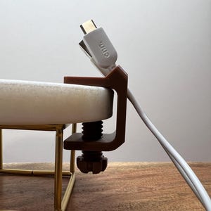 May include: A brown plastic C-clamp cable organizer attached to the edge of a white circular surface. The clamp features a threaded screw mechanism and holds a white USB-C cable with the text "onn." printed on the connector head.