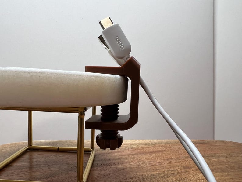 May include: A brown plastic C-clamp cable organizer attached to the edge of a white circular surface. The clamp features a threaded screw mechanism and holds a white USB-C cable with the text "onn." printed on the connector head.
