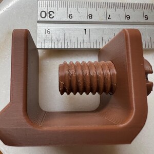 May include: A brown C-clamp with a threaded screw mechanism, positioned next to a metal ruler showing a measurement of approximately 1 inch for the screw diameter. The clamp features a U-shaped frame and a textured surface from 3D printing.