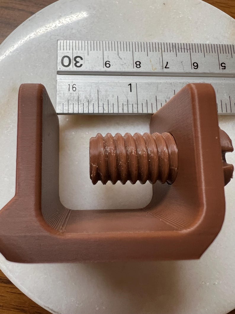May include: A brown C-clamp with a threaded screw mechanism, positioned next to a metal ruler showing a measurement of approximately 1 inch for the screw diameter. The clamp features a U-shaped frame and a textured surface from 3D printing.
